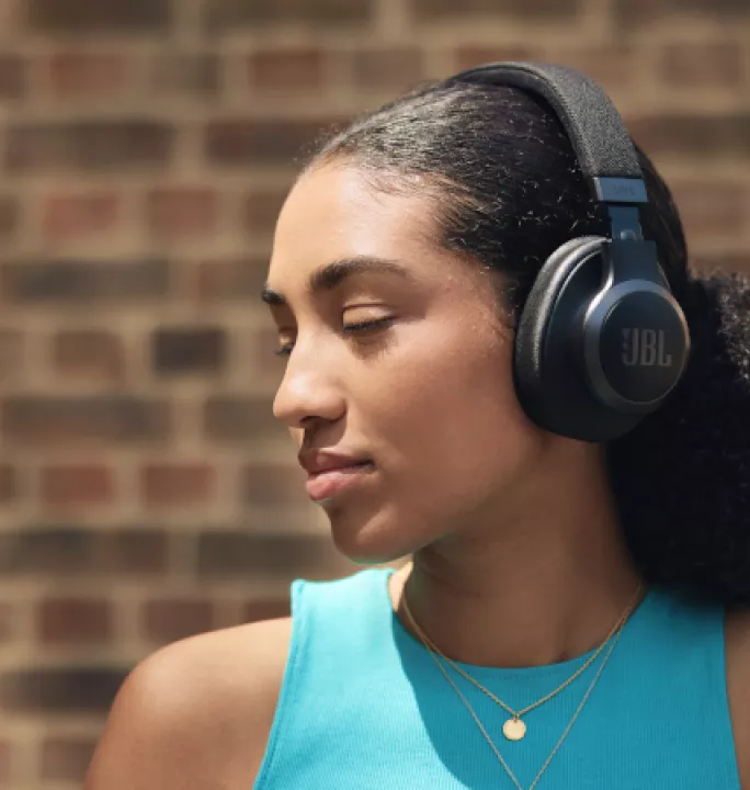 A young woman with large black JBL headphones stands against a brick wall. She appears relaxed with eyes closed, wearing a turquoise tank top and gold necklace.