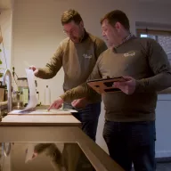 Two men wearing olive-colored "Mijn tuintje" sweaters reviewing documents and using a tablet in a kitchen setting, appearing to discuss garden plans or designs for Bestselect.