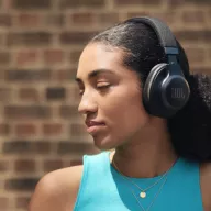 A young woman with large black JBL headphones stands against a brick wall. She appears relaxed with eyes closed, wearing a turquoise tank top and gold necklace.
