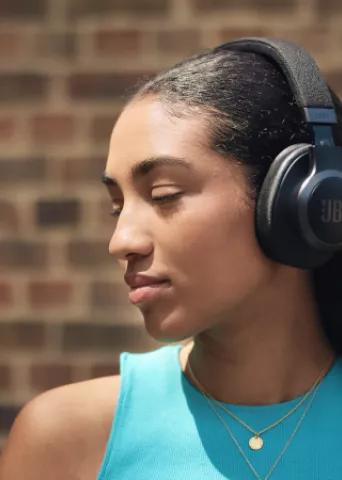 A young woman with large black JBL headphones stands against a brick wall. She appears relaxed with eyes closed, wearing a turquoise tank top and gold necklace.