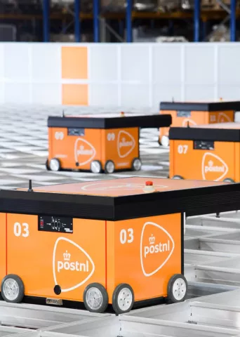 Automated guided vehicles (AGVs) with orange tops and blue bases move efficiently along tracks in a large, modern PostNL fulfillment warehouse, demonstrating advanced logistics automation.