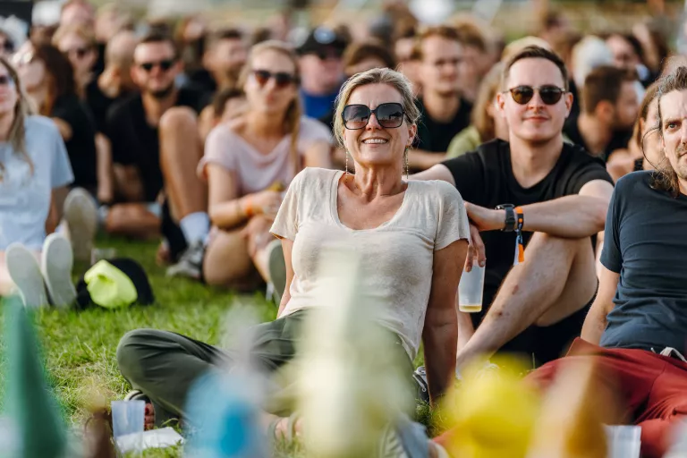Smiling woman from Springbok Agency in sunglasses sits on grass at the outdoor summer event. People around her relax and enjoy the sunny atmosphere, conveying a joyful mood.