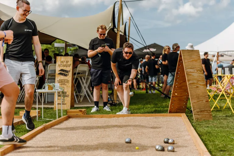 A group of men play pétanque at Springbok's outdoor team event. One is in mid-throw, smiling. Others watch or use their phones. The mood is relaxed and cheerful.