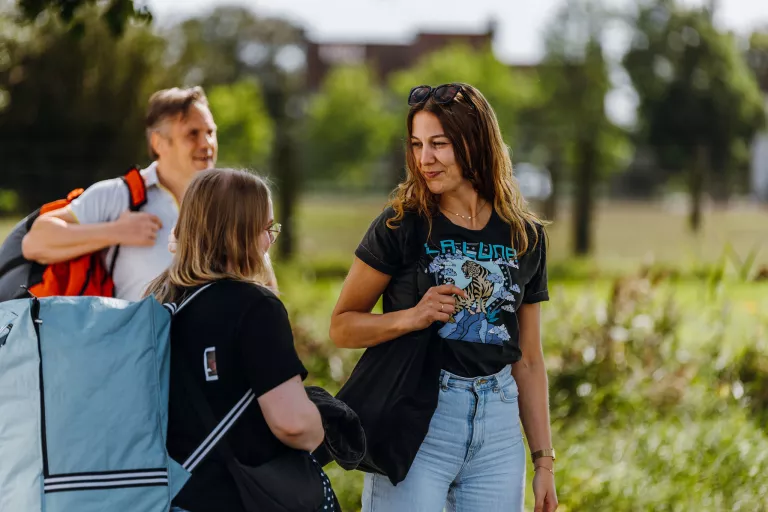 Three people, smiling and chatting outdoors. A woman in a graphic tee is in focus, with another woman and a man carrying a backpack in the background. Bright, sunny day.