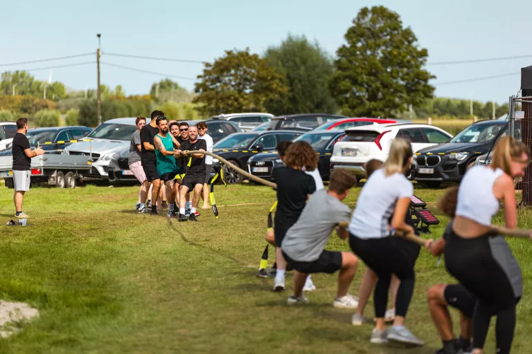 Two teams energetically pull on a rope in a tug-of-war at Springbok's summer team event. They are on a grassy field next to parked cars, conveying a sense of fun and teamwork.