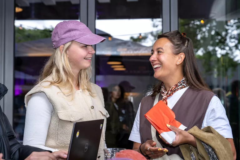 Two women from Springbok Agency smiling and conversing outside. One wears a purple cap and holds a laptop, the other has a scarf and holds a red pouch.