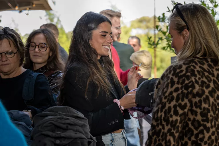 A group of people stand in conversation outdoors. A woman in a black jacket smiles warmly. The atmosphere is casual and friendly.