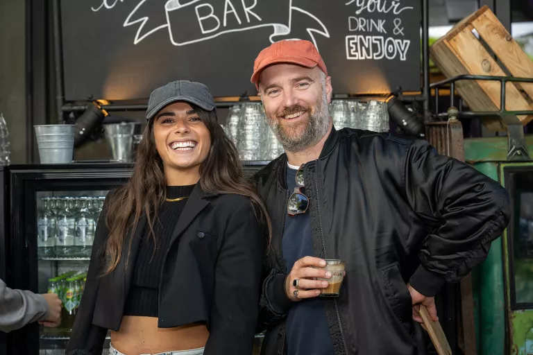 A man and woman from Springbok Agency smiling at an outdoor bar. The woman wears a gray cap, black top, and beige pants. The man wears a red cap and black jacket, holding a drink.