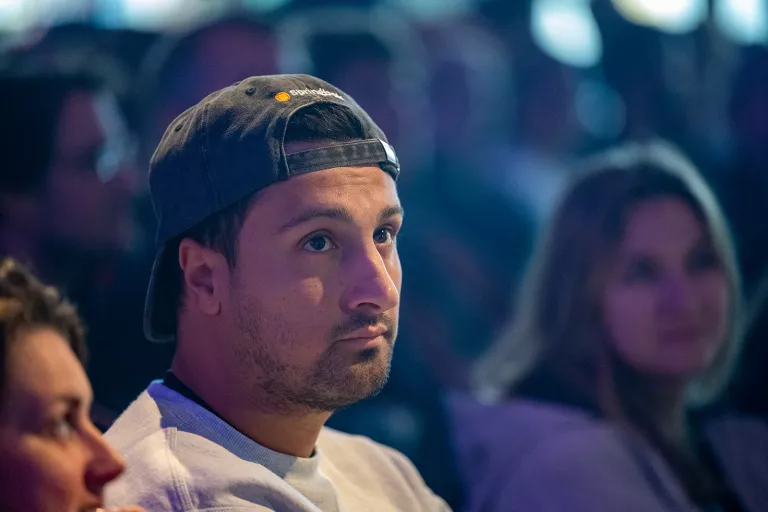 A man wearing a backwards cap and hoodie gazes attentively in a dimly lit room, surrounded by blurred audience members at a Dutch Springbok team event.