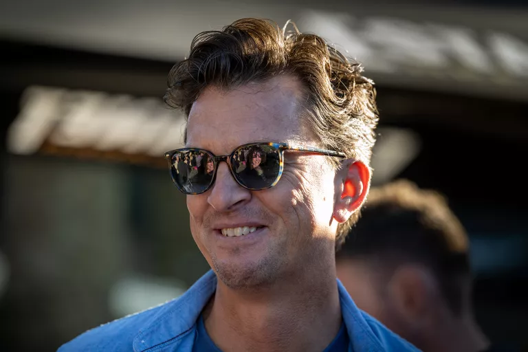 Man from Springbok Agency with curly hair wearing sunglasses and a blue shirt, smiling outdoors. Reflections on his glasses show a crowd. Bright sunlight and relaxed mood.