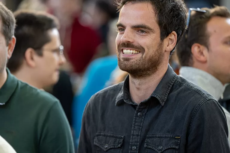 A smiling man in a black denim shirt stands in a crowded room, conveying a cheerful atmosphere. Blurred people in the background enhance the social setting.