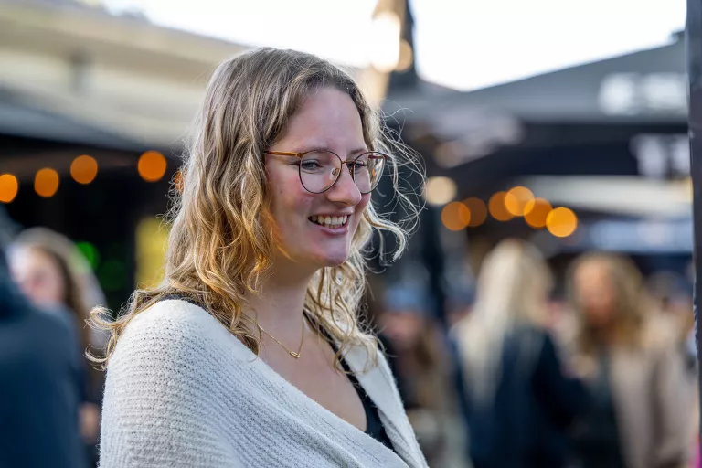 A smiling woman with wavy hair and glasses stands outdoors, wearing a cream sweater. The blurred background shows people and string lights at Springbok's summer team event.