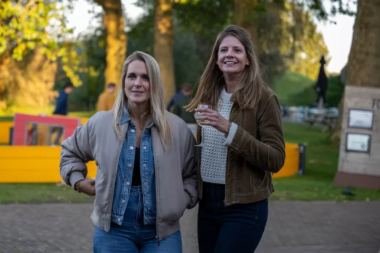 Two women from Springbok Agency standing outdoors, one in a beige jacket and the other in a brown jacket holding a drink. They smile in a sunny park setting with tall trees.