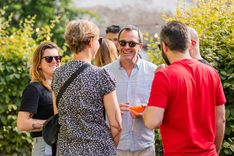 A group of people is enjoying a conversation outdoors, surrounded by greenery. They are smiling and laughing, creating a warm, relaxed atmosphere.