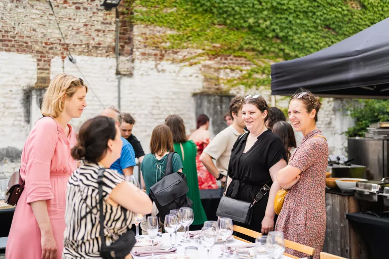 People chatting and smiling by a dining table set with glasses at Springbok's summer barbecue event in Ghent. An ivy-covered brick wall creates a relaxed, casual atmosphere.