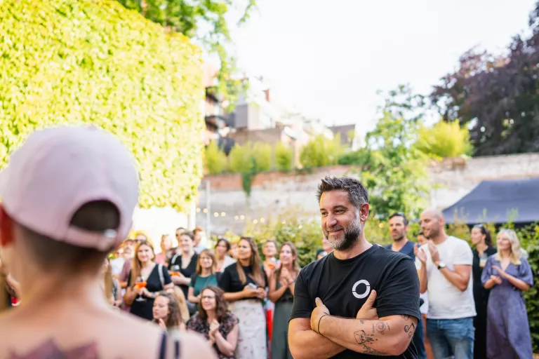 A man in a black T-shirt smiles with arms crossed at Springbok's summer barbecue in Ghent. A woman in a pink cap speaks to a crowd. The mood is cheerful and social.
