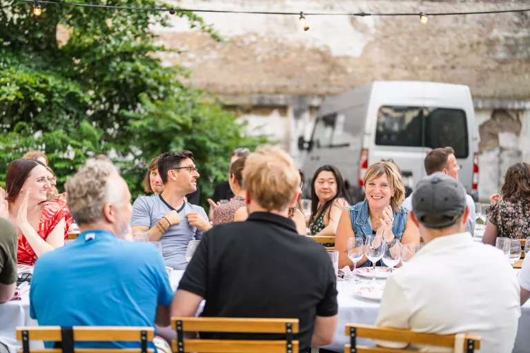 Group of people sitting at Springbok's outdoor summer barbecue in Ghent, chatting and smiling around tables with white tablecloths and wine glasses.