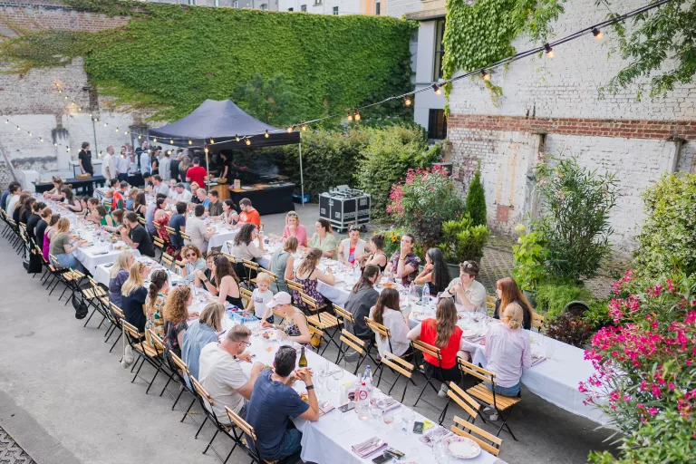 Springbok summer barbecue event in Ghent, with people seated at long tables under string lights in a garden-like setting. Lively atmosphere with greenery and flowers.