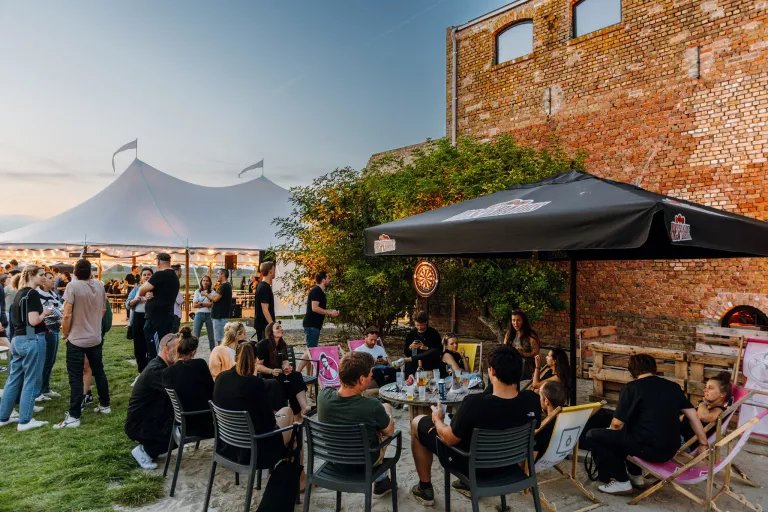  Outdoor Springbok event with people under a canopy and umbrella near a brick wall. A group is seated, some standing, relaxed and socializing. Tent with warm lights in the background.