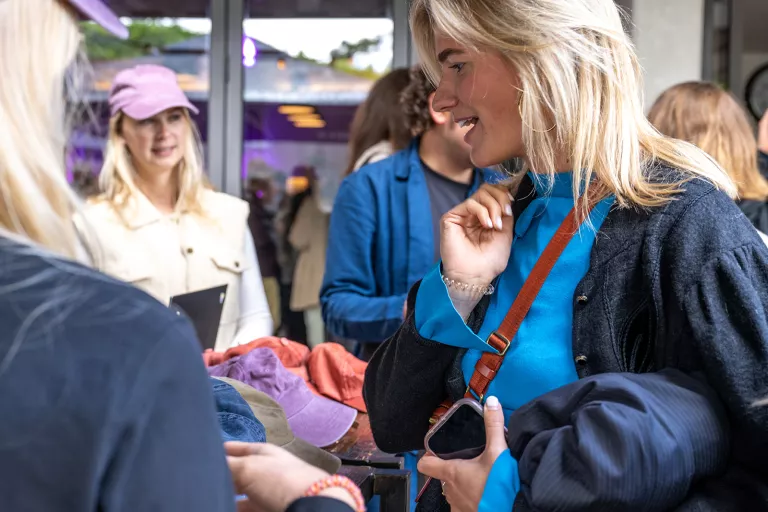 A group of people shopping at a Springbok team event. A smiling woman in a blue top holds a phone and coat, while colorful hats are on display. Relaxed atmosphere.