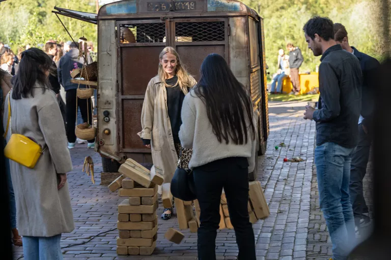 A group of people enjoy a Springbok team event. A woman in a beige coat smiles near a wooden block game that is falling, next to a vintage van and baskets.