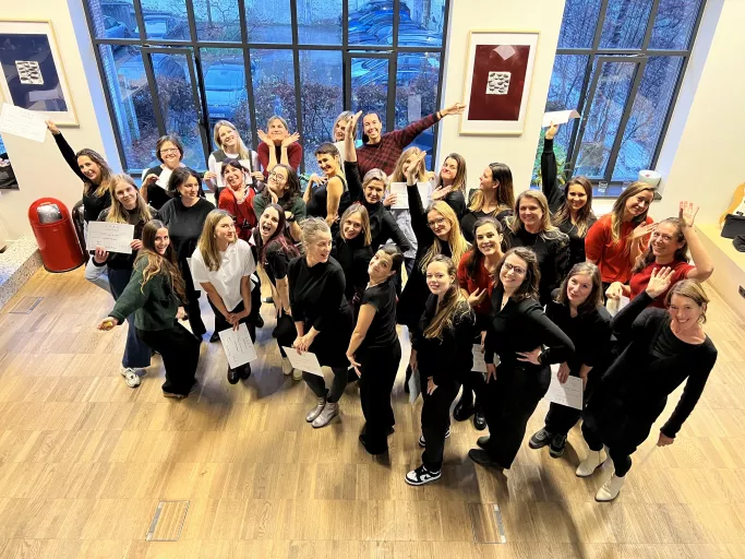Twenty-nine female Springbok team members stand in a sunlit room with large windows, smiling and celebrating. Some hold certificates. The mood is joyful and lively.