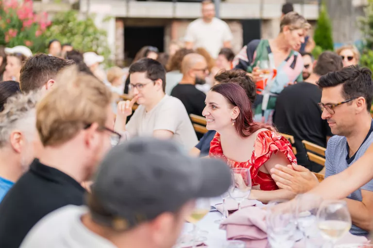 A Springbok summer barbecue team event with people seated at a long table. Tatiana, part of Springbok, is wearing a red dress and smiles while chatting. The atmosphere is joyful and social.