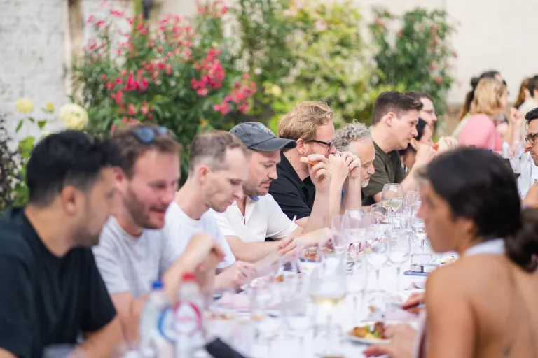 A group of people at the Springbok barbecue event sit at a long outdoor table, eating and talking, with plates, glasses, and greenery visible in the background.