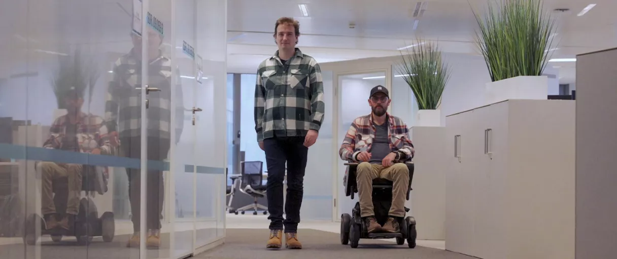 Header image for an ENGIE accessbility article with two men in an office hallway, one standing and one sitting in a motorized wheelchair; both are wearing plaid shirts and boots.