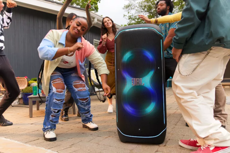 A group of young Gen Z people dances energetically outdoors near a large illuminated JBL speaker. The scene is lively and colorful, capturing joy and celebration.