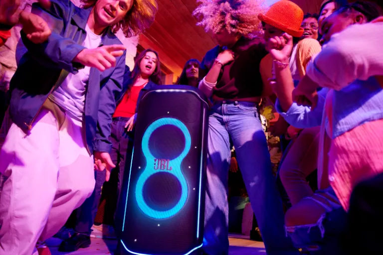 Party scene with diverse group of Gen Z people dancing inside around a glowing JBL speaker. We see vibrant lighting, an energetic atmosphere, colorful outfits and joyful expressions.
