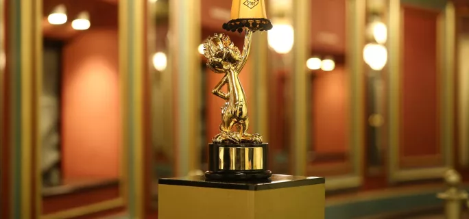A golden Loekie Award trophy featuring a stylized dog holding a small lampshade with the word "Ster" on it, placed on a black and gold pedestal indoors with a blurred background and warm lighting.